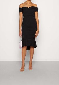 Vestido negro off-the-shoulder con una silueta ajustada, dobladillo acampanado y tejido texturizado. Combinado con sandalias transparentes y un pequeño bolso rosa.