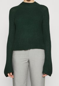 Pull en maille vert foncé avec un col montant et des manches larges, présentant un motif côtelé texturé. Associé à un pantalon ajusté gris clair.