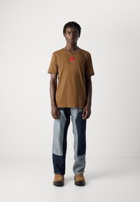 Ung mann står iført en brun t-skjorte med et rødt logo-patch, totonet lappesydd jeans, beige støvler og et sølvkjede.