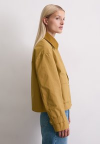 Mosterdgele jas met een corduroy kraag, twee voorkwarten en knoopsluitingen. Gekozen in combinatie met blauwe spijkerbroeken, zijaanzicht.