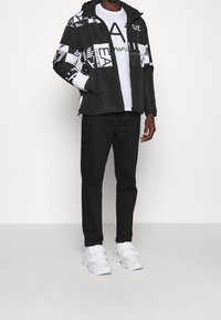 Svart och vit mönstrad jacka med huva, i kombination med en vit t-shirt och svarta byxor, fulländad med vita sneakers.