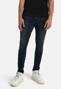 Jeans de mezclilla oscura ajustados, con una textura suave, un ligero desgastado y un diseño clásico de cinco bolsillos, combinados con zapatillas altas blancas.