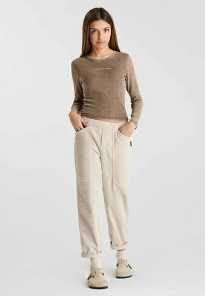 Jeune femme debout, les mains dans les poches, portant un haut marron à manches longues, un pantalon beige en velours côtelé et des chaussures beige sans lacets avec des chaussettes.