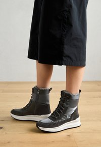 Botas de tobillo grises y negras con un cuello forrado de piel, cierre de cremallera y materiales texturizados. Presenta una suela blanca gruesa y paneles en contraste.