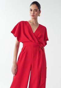 Punane ülikond lühikeste lendavate varrukatega, V-kaelusega ja sidumisega vöökohal. Sile kangas, vabalt langev lõige ja laiad sääred.