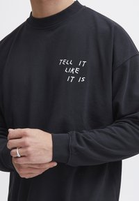 Black sweatshirt with a ribbed crew neck, featuring white text "TELL IT LIKE IT IS" printed on the left side. Soft fabric and long sleeves.