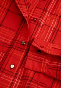 Czerwona flanelowa koszula w kratę z czarnymi i pomarańczowymi liniami; posiada metalowe guziki, kieszeń na piersi oraz teksturowany wzór materiału.
