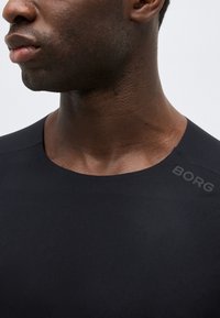 Κοντινό πλάνο του κάτω μέρους του προσώπου και του λαιμού ενός ατόμου που φοράει μαύρο πουκάμισο με την ένδειξη «BORG» τυπωμένη κοντά στον γιακά στη δεξιά πλευρά.