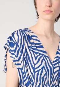Robe à v-neck rayée bleue et blanche en tissu léger, avec des manches courtes à liens et une coupe décontractée. Tatouage notable visible.