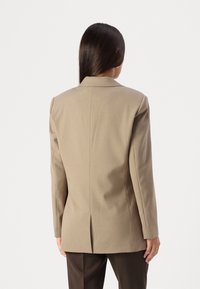 Blazer beige cintré avec des épaules structurées, une fente au dos et des manches longues. Tissu lisse avec une finition soignée. Associé à un pantalon marron.