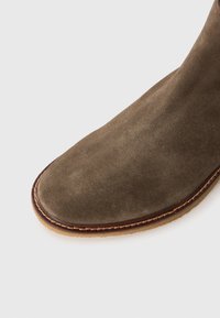 Primer plano de la punta de una bota de tobillo de ante marrón con costuras detalladas y suela de goma de color marrón claro sobre fondo blanco.