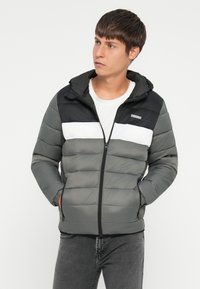 Νεαρός άνδρας φορώντας ένα γκρι, μαύρο και άσπρο χρωματιστό τζάκετ με κουκούλα, με τα χέρια στις τσέπες, κοιτάζοντας στο πλάι.