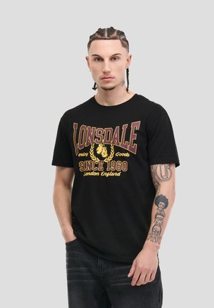 Young man with braided hair wearing a black Lonsdale t-shirt and dark jeans, showing tattoos on his left forearm and hand in neutral pose.