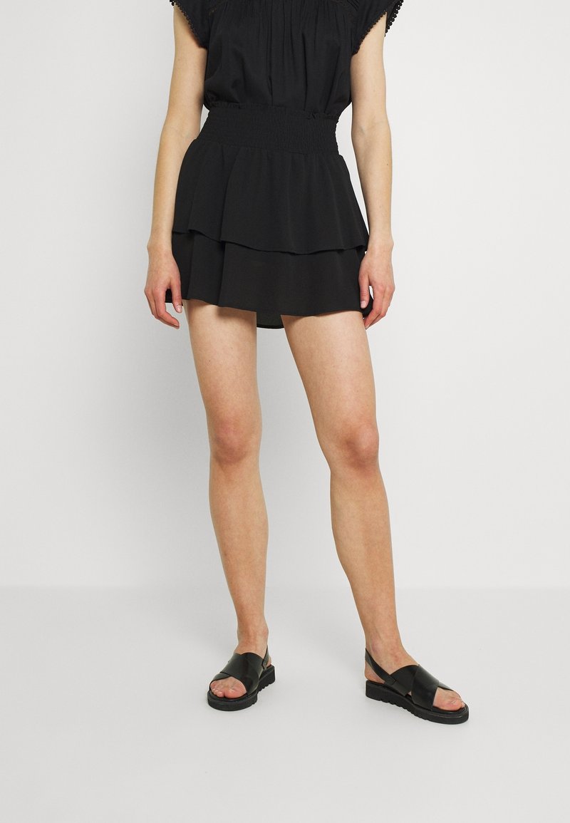 Femme portant une mini-jupe noire à volants avec une taille smockée et des sandales noires en cuir à bout ouvert, debout devant un fond clair uni.