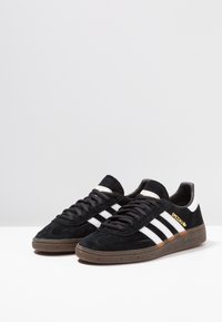 Zapatillas de ante negras con diseño de tres rayas blancas, parte superior texturizada y acento dorado "Spezial". Suela de goma con detalles marrones.