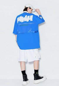 Camisa azul de gran tamaño con un gráfico blanco, detalle con cremallera y texto verde. Combinada con pantalones cortos cargo blancos y botas negras.