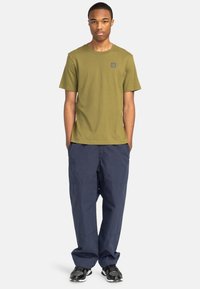 Olivgrünes Baumwoll-T-Shirt mit kleinem Logo auf der Brust, kombiniert mit lockeren navyblauen Cargo-Hosen und schwarzen Sportschuhen.