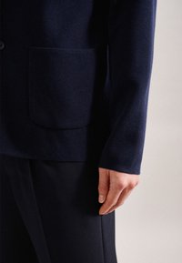 Navyblå texturerad jacka med bröstficka och åtsittande ärm, ihop med mörka byxor. Fokusera på tyg- och design detaljer.