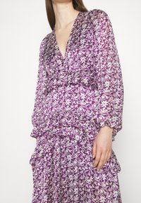 Robe florale violette avec un design à manches longues bouffantes, volants superposés et un décolleté en V profond. Fabriquée en tissu léger et translucide.
