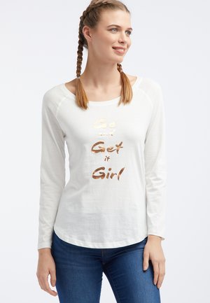 Kobieta z plecionymi włosami, ubrana w białą koszulę z długim rękawem z złotym napisem „Go and Get it Girl” oraz niebieskie dżinsy.
