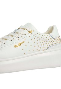 Hvite skinn-sneakers med gullnagler, flate lisser og tykk hvit såle, med merkevarenavnet i gull på siden.