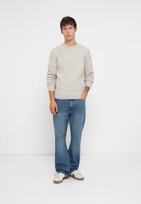 Un pull en tricot gris clair avec un col rond, porté avec un jean en denim bleu. Le modèle se tient les mains dans les poches, portant des baskets blanches.