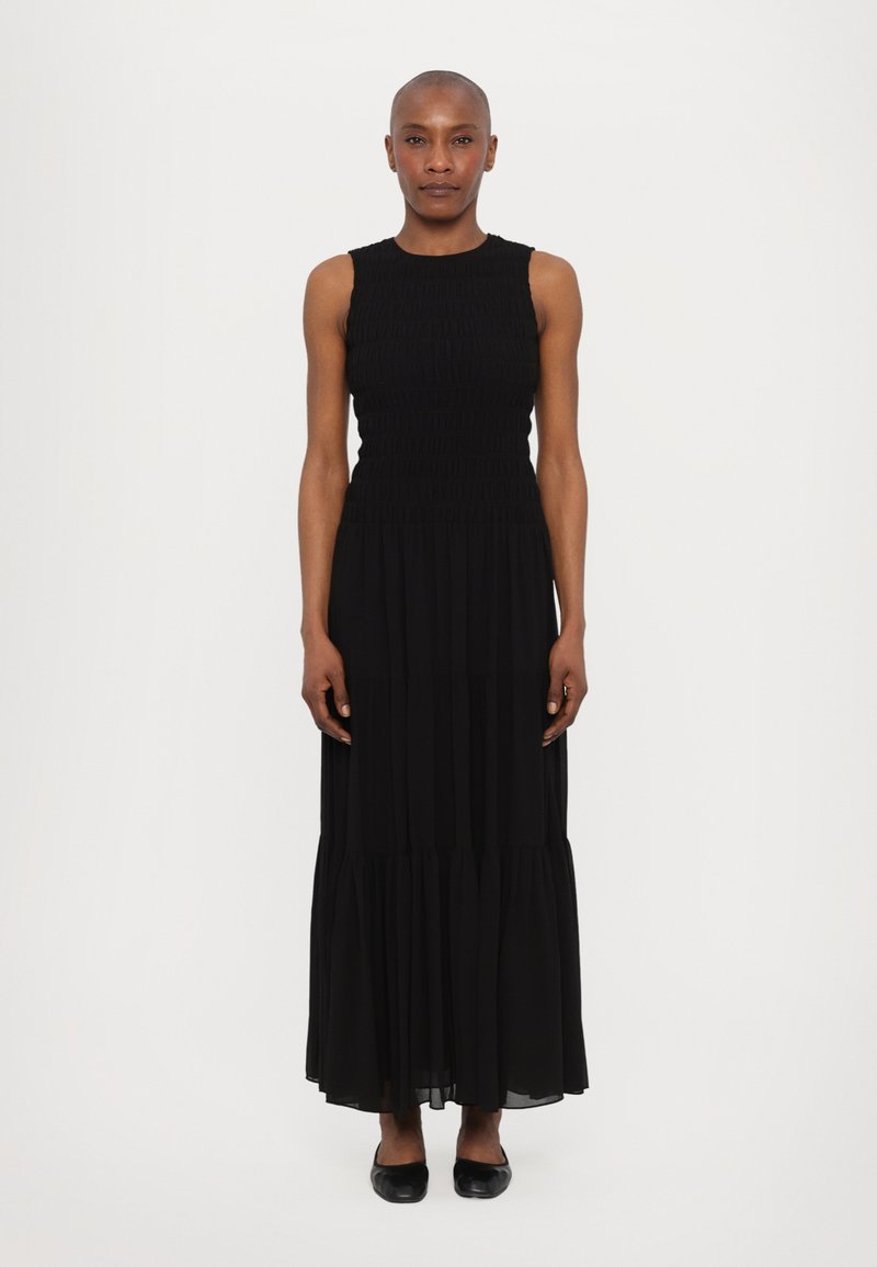 Rochie maxi neagră cu un corset texturat, fără mâneci, și o fustă învolburată și etajată. Țesătură netedă cu un finisaj moale. Pantofi negri fără toc.