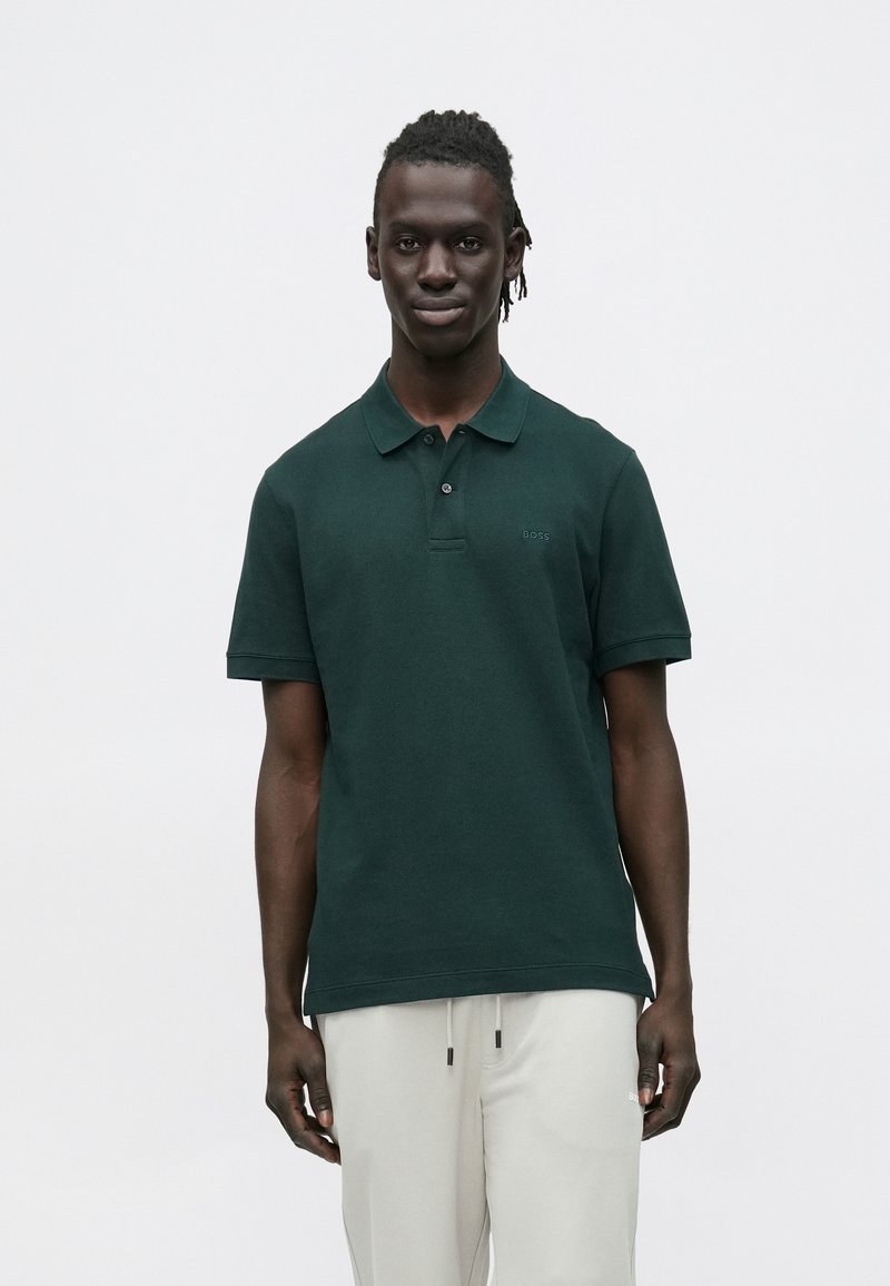Ciemnozielona koszulka polo wykonana z teksturowanej tkaniny, z zapinanym na guziki karczkiem i małym logo na lewej piersi. Połączona z jasnymi spodniami.