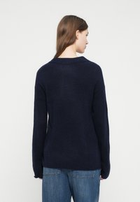 Μπλε ναυτικό πουλόβερ με ευρύ ντεκολτέ, μακριά μανίκια και ριμπ μανσέτες, κατασκευασμένο από απαλό υφασμάτινο ύφασμα με υφή. Συνδυάζεται με τζιν παντελόνι ανοιχτό μπλε.