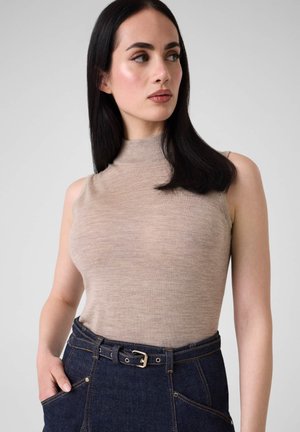 Femme aux longs cheveux noirs portant un col roulé beige sans manches et un jean taille haute bleu foncé avec ceinture, regardant vers sa gauche.