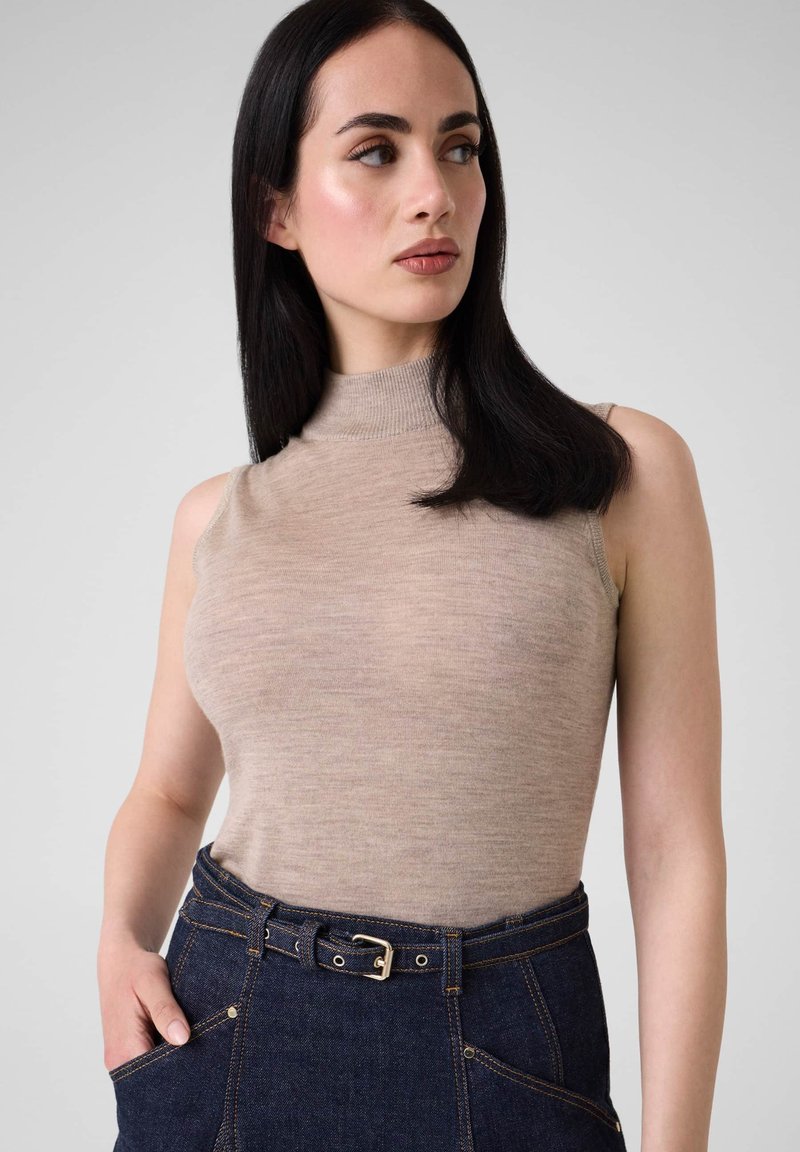Femme aux longs cheveux noirs portant un col roulé beige sans manches et un jean taille haute bleu foncé avec ceinture, regardant vers sa gauche.