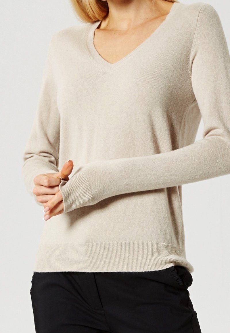 Femme portant un pull beige clair à col en V, manches longues avec poignets et ourlet côtelés, associé à un pantalon noir.