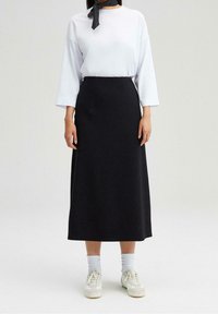Sort, gulvlange nederdel med en glat tekstur og vinklet sømdesign, parret med en hvid langærmet t-shirt og hvide sneakers.