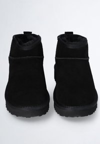 Un par de botines de ante negro con forro de piel y suelas de goma gruesas, mostrados de frente sobre un fondo blanco.