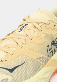 Žlto-béžová bežecká obuv Hoka so šnúrkami, sieťovinovým materiálom a viditeľným logom na strane a jazyku.