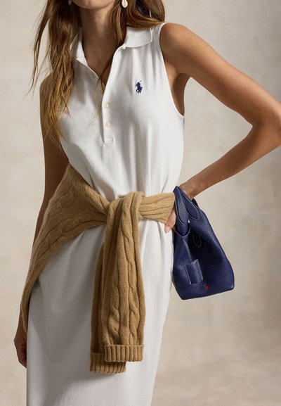 Femme portant une robe polo blanche sans manches avec un pull beige noué autour de la taille, tenant un petit sac à main bleu marine.