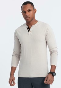 Vaaleanbeige pitkähihaisen henley-paidan, joka on valmistettu pehmeästä kankaasta. Paidassa on musta nappilista ja kolme nappia, ja se on yhdistetty tummiin housuihin.