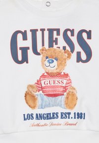 Weißes Sweatshirt mit einem Grafikmotiv eines Teddybären in einem rot gestreiften Hemd mit der Aufschrift "GUESS." Der Text enthält "LOS ANGELES EST. 1981" und "Authentic Denim Brand."