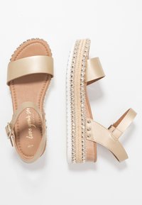 Beige offene Plattformsandalen mit metallischen Nieten, verstellbaren Knöchelriemen und gepolsterten Innensohlen vor weißem Hintergrund.