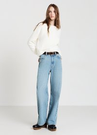 Junge Frau in einem weißen Pullover, hellblauen, hoch taillierten Jeans mit brauem Gürtel und schwarzen, mit Nieten besetzten Holzschuhen, steht vor weißem Hintergrund.