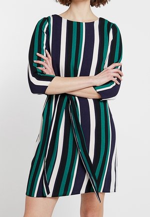 Femme portant une robe mi-longue à rayures verticales bleu marine, vert, blanc et noir, les bras croisés sur la poitrine.