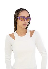 Occhiali da sole rettangolari viola con lenti gialle e finitura opaca. Indossati con una maglietta bianca a maniche lunghe con scollatura a coste.
