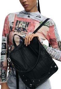 Donna che indossa una maglietta a maniche lunghe multicolore e jeans chiari, mentre tiene una grande borsa in pelle nera con borchie metalliche e dettagli in cerniera.