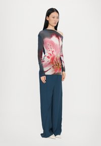Langarmshirt mit floralen Mustern in Pink und Rot, kombiniert mit lockeren, marineblauen Hosen und spitzen, weißen Schuhen. Aus glattem, leichtem Stoff.
