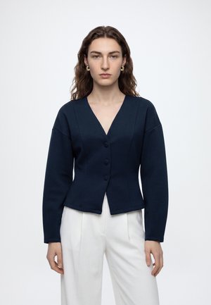 Femme aux cheveux bruns ondulés, portant des boucles d’oreilles en or, une veste bleu marine boutonnée à col en V, et un pantalon blanc sur un fond blanc uni.