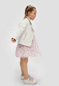 Weiße Kunstlederjacke über einem rosa Blumenkleid mit blauen und lila Mustern. Beige Sneakers mit pastellfarbenen Riemen. Seitenansicht.