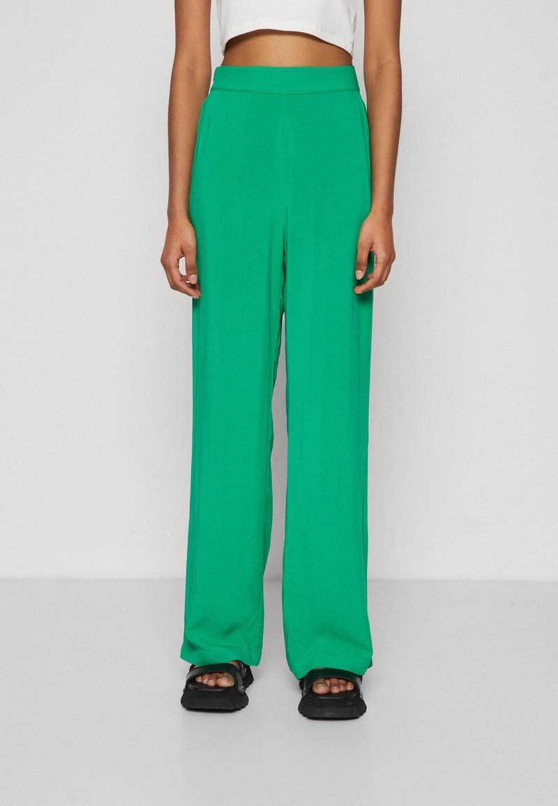 Pantalons larges verts en tissu léger, avec une taille haute et une texture lisse, associés à des sandales noires.
