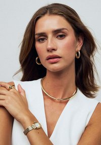 Guld smycken som inkluderar en tjock hoopörhänge, en elegant choker och en armbandsur med silverarmband, placerade mot en vit topp.