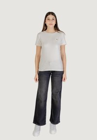 T-shirt grigia in cotone a maniche corte, con un piccolo logo sul petto, abbinata a jeans ampi in denim scuro e sneakers bianche.