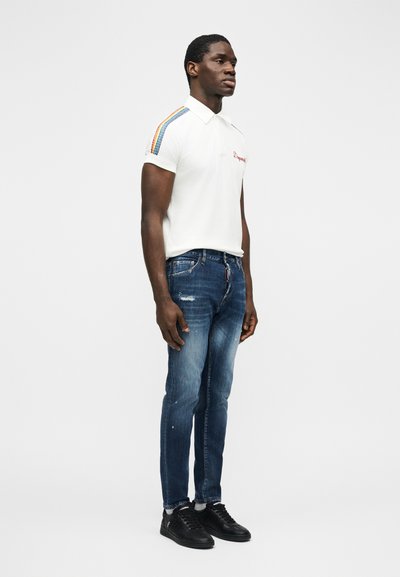 Homme debout portant un polo blanc avec des rayures arc-en-ciel sur les épaules, un jean bleu déchiré et des baskets noires, sur fond blanc.